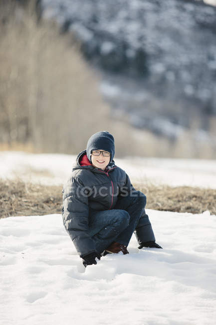 Ragazzo in cappotto invernale e cappello di lana accovacciato nella neve . — Foto stock