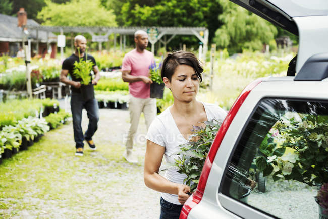 Woman loading flowers into trunk of car parked at garden center with men carrying plants in background. — Stock Photo
