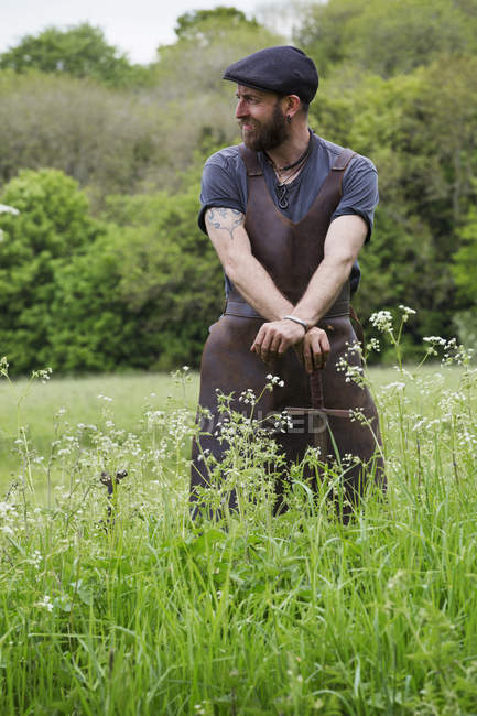 Man in leather apron standing in green field and leaning on vintage sword. — Stock Photo