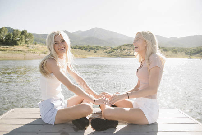 Две блондинки-девочки сидят на причале у озера и держатся за руки . — стоковое фото