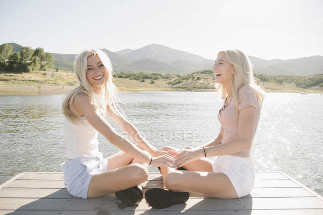 Two blonde teenage girls sitting on lake jetty and holding hands. — Stock Photo