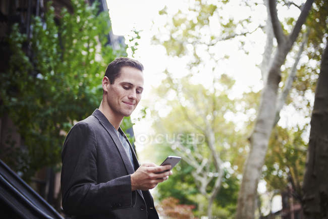 Молодой бизнесмен стоит на городской улице и смотрит вниз на смартфон . — стоковое фото