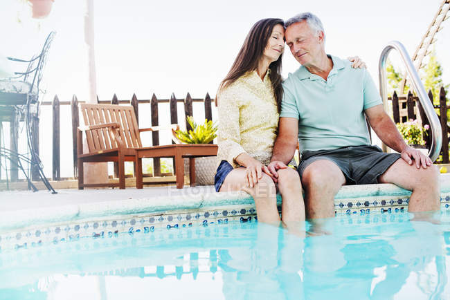 Senior couple sitting head to head on edge of swimming pool. — Stock Photo