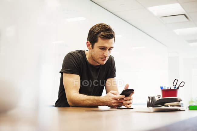 Junger Mann sitzt im Büroraum, lehnt sich auf Schreibtisch und blickt auf Smartphone herab. — Stockfoto