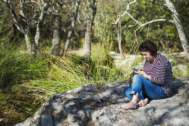 Женщина сидит на скалах у реки и использует смартфон . — стоковое фото