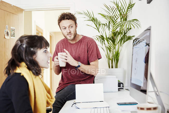 Жінка працює за столом і чоловік сидить на столі з чашкою кави, розмовляє і дивиться на екран комп'ютера . — стокове фото