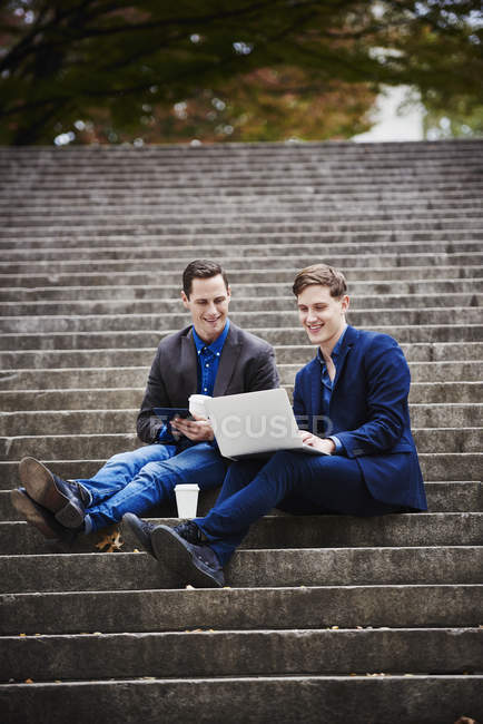 Два молодых человека сидят на ступеньках в городе и используют ноутбук вместе . — стоковое фото