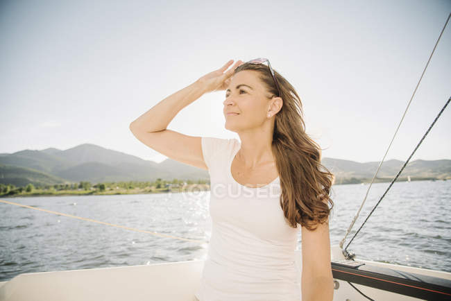 Frau mit langen braunen Haaren steht auf Segelboot und Sonnenbrille am See. — Stockfoto