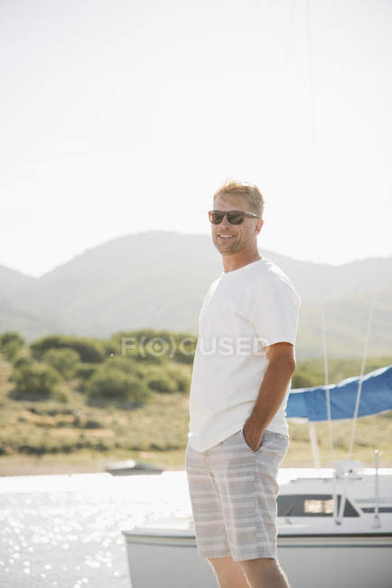 Blond man wearing sunglasses and standing on lake jetty by sailboat. — Stock Photo
