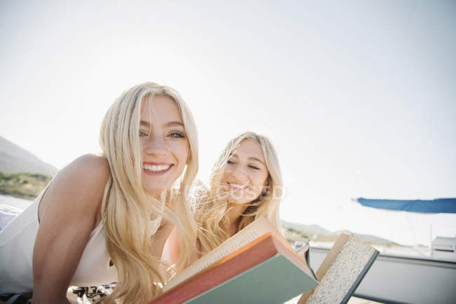Zwei blonde Teenager-Mädchen, die mit Büchern auf dem Steg liegen, lächelnd in die Kamera schauen. — Stockfoto
