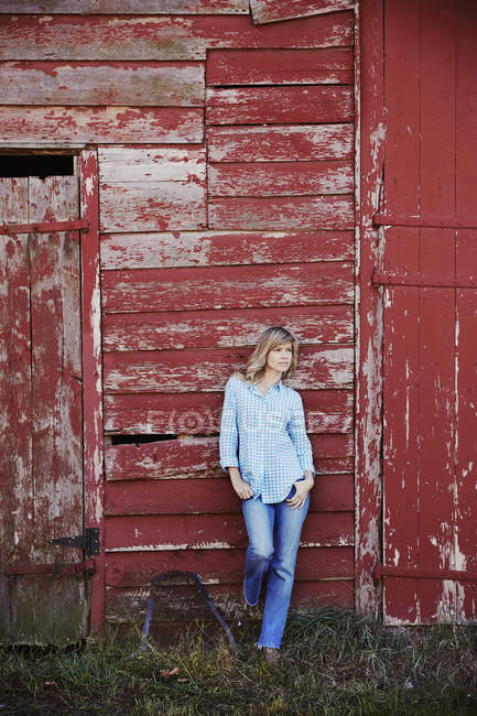 Mid adult woman in jeans leaning against red painted barn building. — Stock Photo