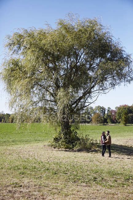 Young couple standing and looking at each other by tree in countryside field. — Stock Photo