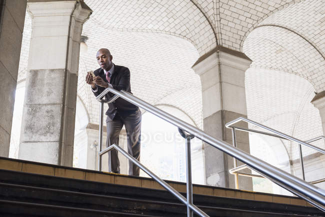 Зрелый бизнесмен в костюме стоит у лестницы под арочным потолком и держит смартфон . — стоковое фото
