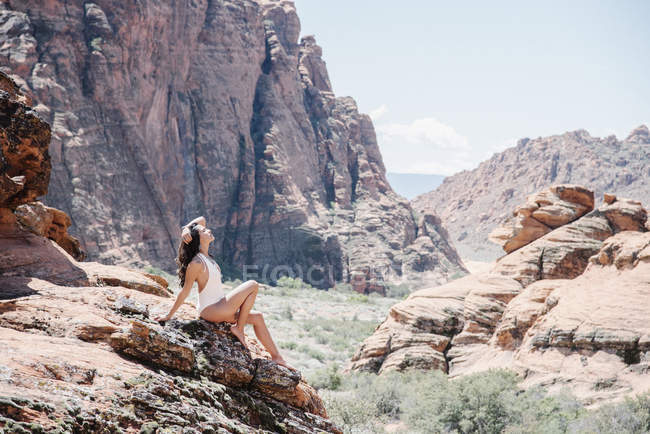 Young woman in white swimsuit resting on rocks in canyon valley with arm raised. — Stock Photo