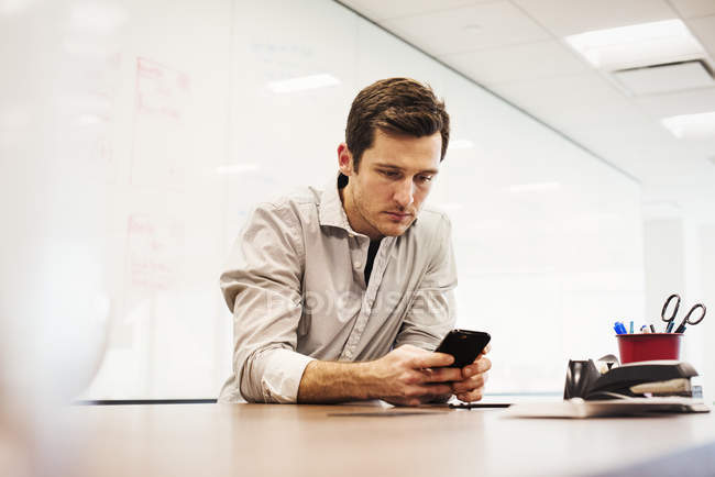 Young man sitting in office room, leaning on desk and looking down at smartphone. — Stock Photo