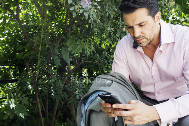 Empresario en traje gris y camisa rosa sentado al aire libre y usando el teléfono . - foto de stock