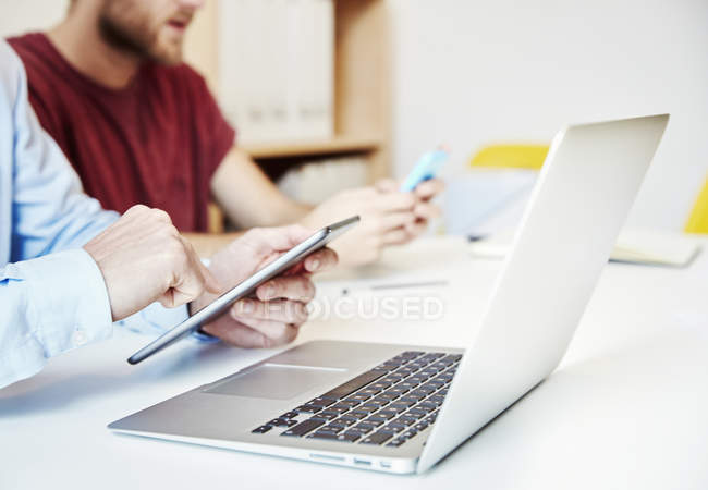 Двое мужчин с ноутбуком, цифровым планшетом и смартфоном сидят за рабочим столом . — стоковое фото