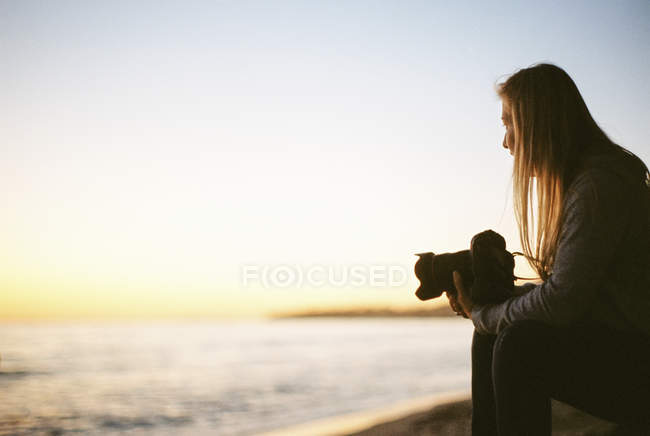 Vue latérale d'une femme aux cheveux blonds assise sur une plage de sable et tenant une caméra . — Photo de stock