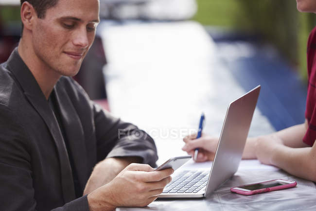 Jovem sentado à mesa ao ar livre com mulher e laptop aberto e olhando para baixo no smartphone . — Fotografia de Stock