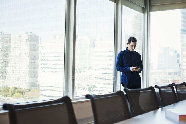 Молодой человек стоит рядом со столом в конференц-зале и смотрит на смартфон . — стоковое фото