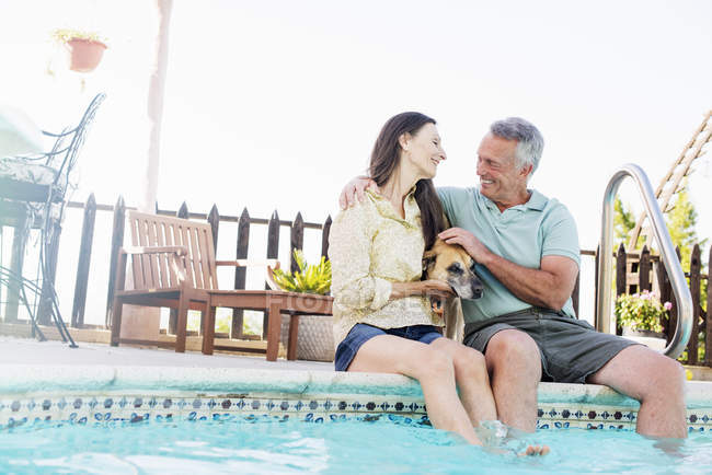 Seniorenpaar sitzt am Rand des Swimmingpools und streichelt Hund dazwischen. — Stockfoto
