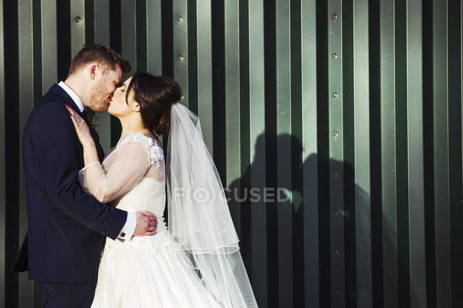 Bride and bridegroom kissing in front of green corrugated metal wall, side view. — Stock Photo