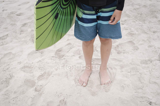 Cropped view of boy standing on sandy beach and holding bodyboard. — Stock Photo