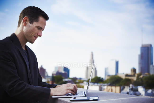 Jovem de pé na construção do telhado e trabalhando com laptop com smartphone ao lado . — Fotografia de Stock