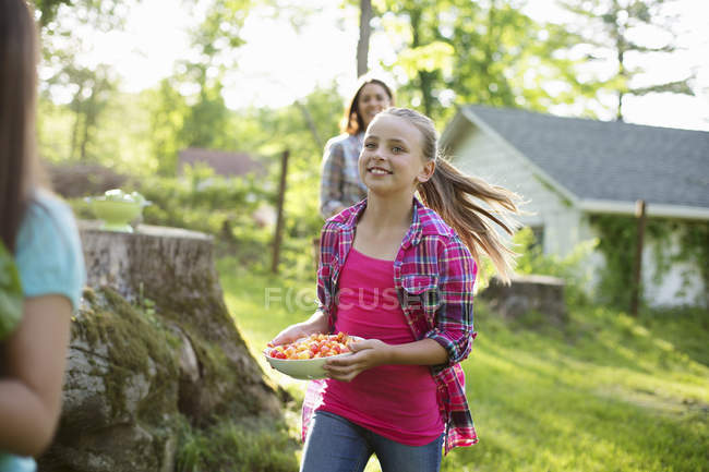 Pre-adolescent girl carrying bowl of cherries on farmhouse lawn with sisters. — Stock Photo