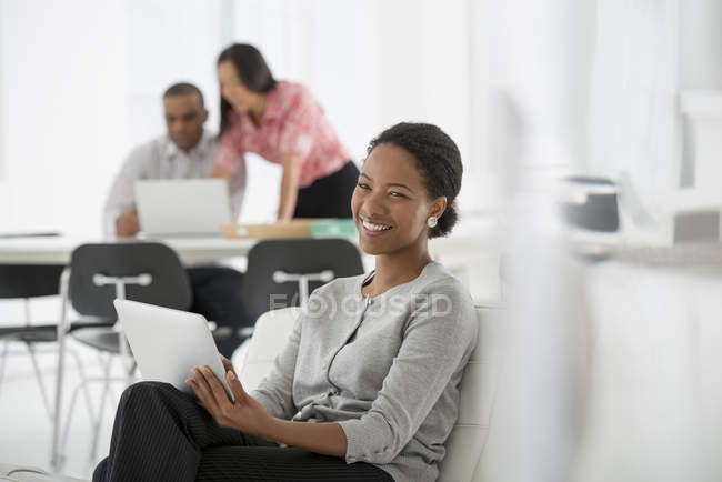 Woman holding digital tablet while sitting on sofa in office with coworkers using laptop in background. — Stock Photo