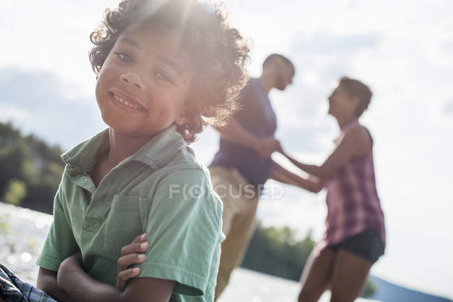 Età elementare ragazzo con le braccia incrociate seduto sul molo del lago soleggiato con i genitori . — Foto stock