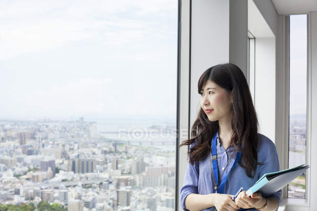 Jeune femme d'affaires tenant des dossiers et regardant par la fenêtre dans un immeuble de bureaux . — Photo de stock