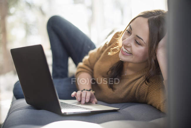 Giovane donna allegra sdraiata sul divano e utilizzando il computer portatile . — Foto stock