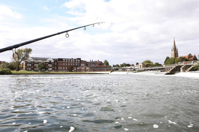 Fishing rod against water by weir and bridge of town in England. — Stock Photo