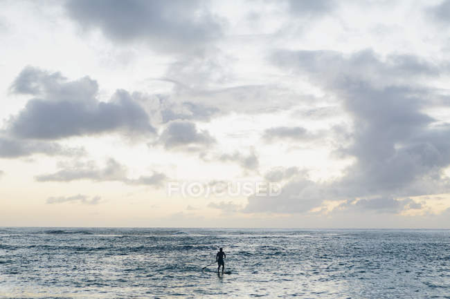 Mann im Stand Up Paddling in ruhigem Wasser in der Dämmerung unter malerischer Wolkenlandschaft. — Stockfoto