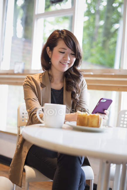 Giovane donna sorridente seduta a tavola con tazza e fetta di torta e guardando smartphone . — Foto stock