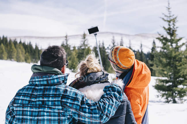 Three people in skiing gear posing for selfie with selfie stick in snowy mountains. — Stock Photo