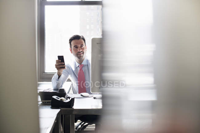 Geschäftsmann hält Smartphone während er im Büro am Computermonitor sitzt. — Stockfoto