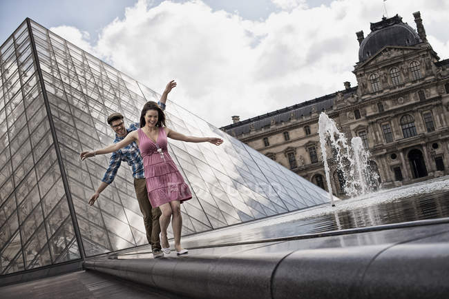Mid adult couple balancing on fountain in courtyard of Louvre museum by glass pyramid in Paris, France. — Stock Photo