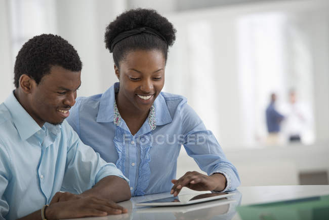 Joven hombre y mujer sentados uno al lado del otro y usando tableta digital en la oficina . - foto de stock