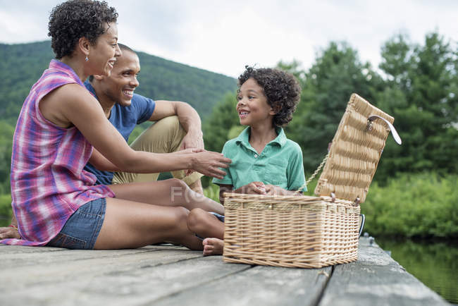 Family with elementary age boy sitting on pier with picnic basket. — Stock Photo