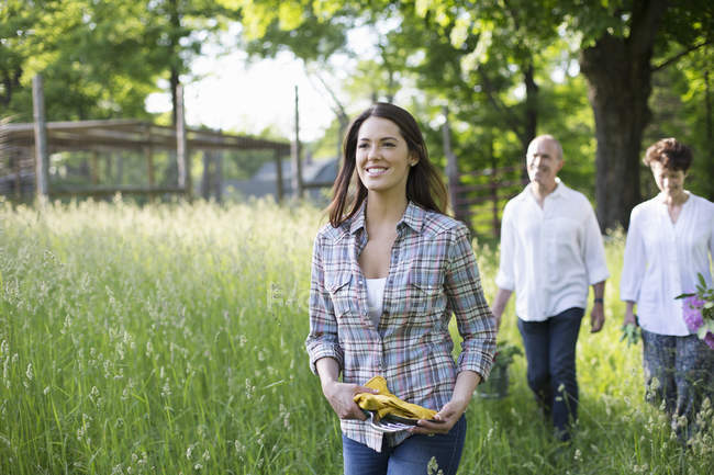 Reifes Paar und junge Frau mit Schutzhandschuhen gehen auf Bauernhof durch langes Gras. — Stockfoto