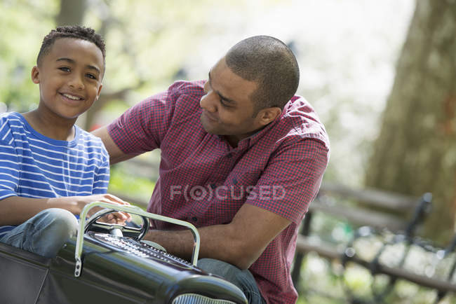Padre ayudando a niño montar a la antigua coche de juguete vendedor ambulante en Sunny Park . - foto de stock