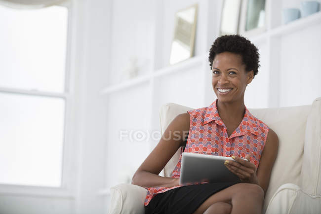 Mulher sorridente sentada no sofá branco e segurando tablet digital . — Fotografia de Stock