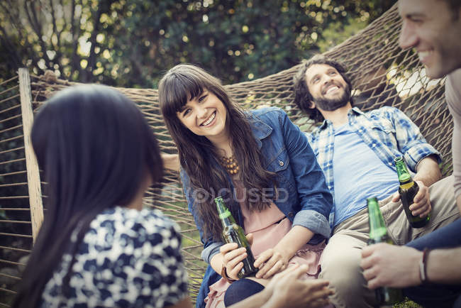 Group of cheerful friends lounging in hammock in garden and drinking beer. — Stock Photo