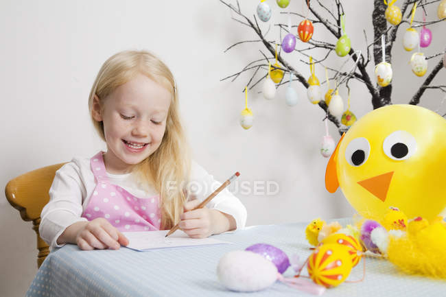 Elementary idade loira menina sentada à mesa e desenho com lápis com ovos de Páscoa decoração no quarto . — Fotografia de Stock