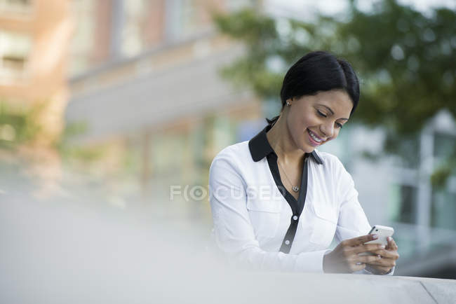 Frau in weißer Jacke checkt Handy, während sie sich an Brüstung in der Stadt lehnt. — Stockfoto