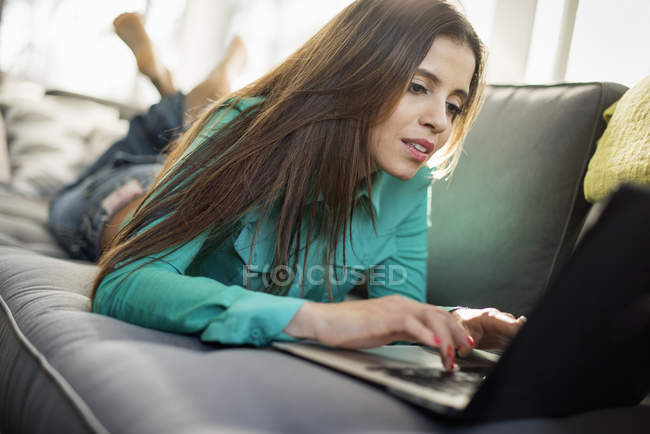 Langhaarige junge Frau liegt auf Sofa und benutzt Laptop. — Stockfoto