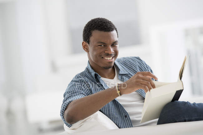 Молодий чоловік сидить з книгою і посміхається в світло-білій кімнаті . — стокове фото