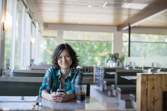 Woman in checked shirt sitting at table, smiling and holding smartphone in diner. — Stock Photo